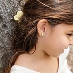 Young girl with a flower in her hair and art palette paint earrings, standing against a natural background.