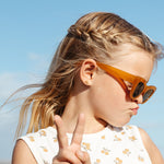 Young girl wearing sunglasses and a white dress with a floral pattern, standing against a clear blue sky.