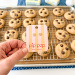 Hand holding a 'pip pop' card in front of a tray of cookies with chocolate chips.