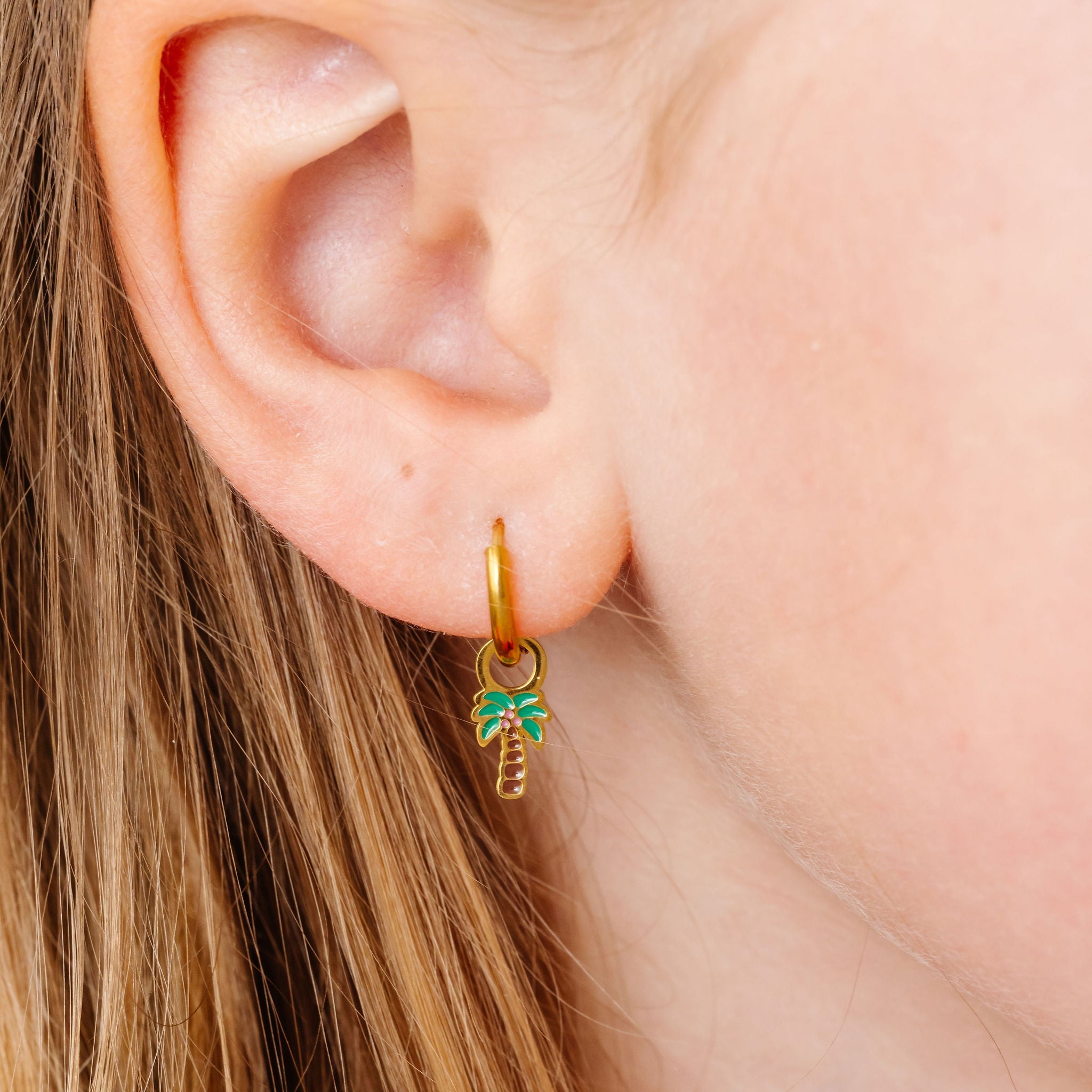 Gold hoop earring with a palm tree design worn by a person with blonde hair.