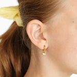 Close-up of a person wearing lemo gold charm earrings with a plain background